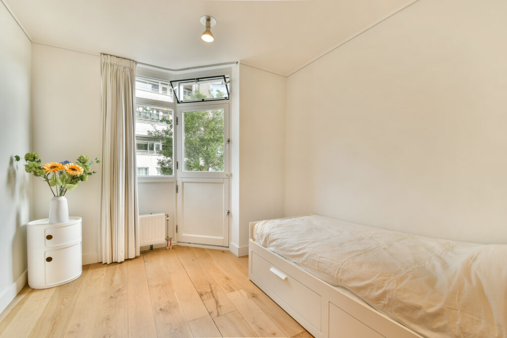 A serene bedroom showcasing minimalist design, featuring a bed, wooden flooring, and large windows that invite natural light, complemented by a vase of flowers.