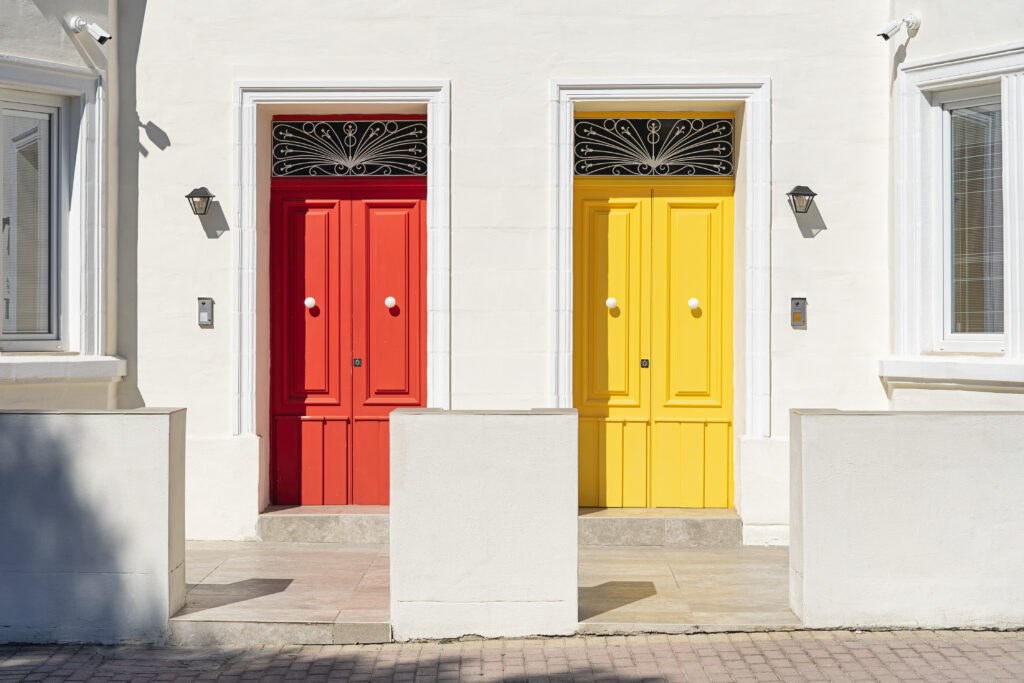Red and yellow door side by side on white building facade, basking in sunlight.